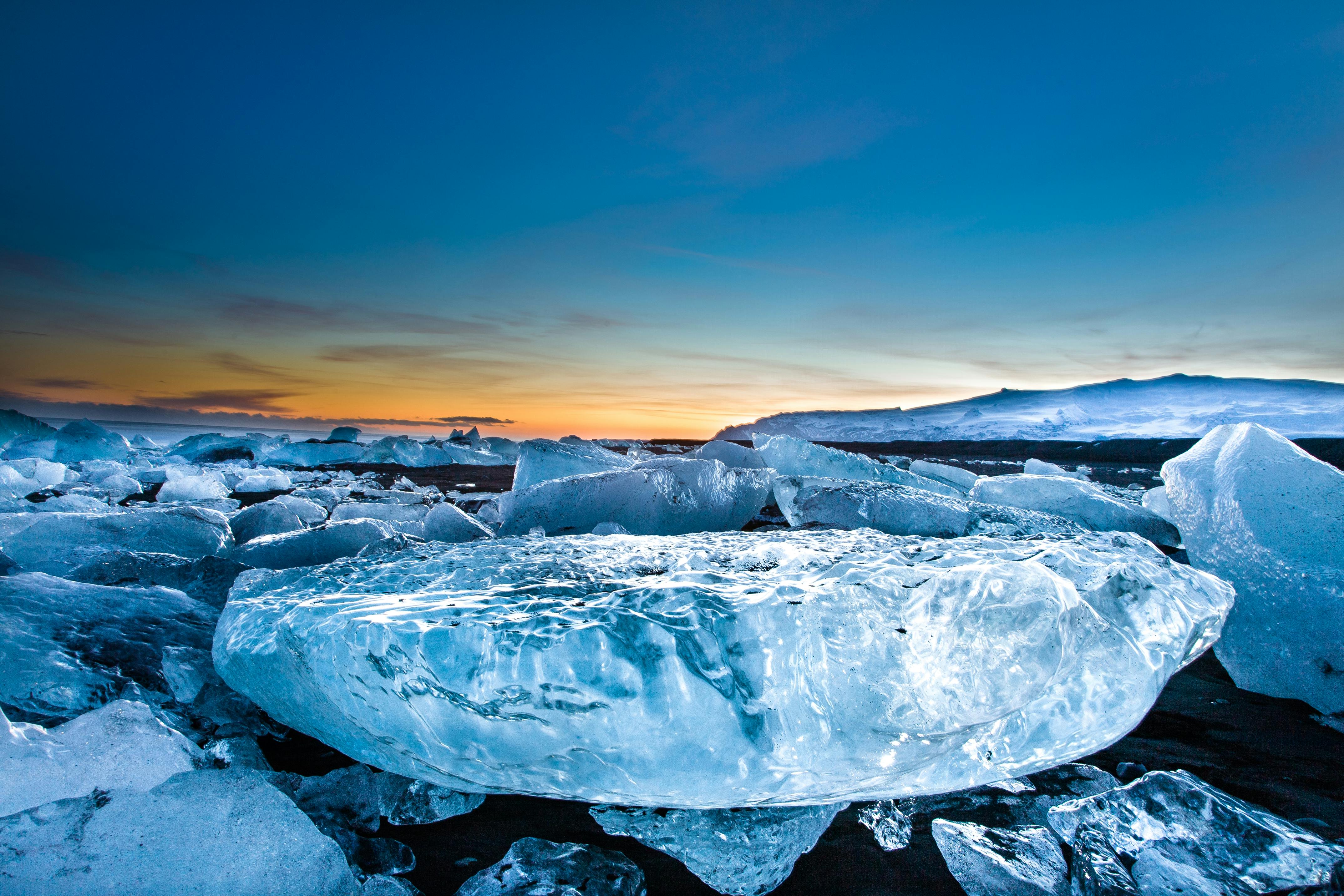 Gros morceaux de glace éparpillés sur un rivage de sable noir sous un ciel crépusculaire, le soleil se couchant derrière des montagnes lointaines.