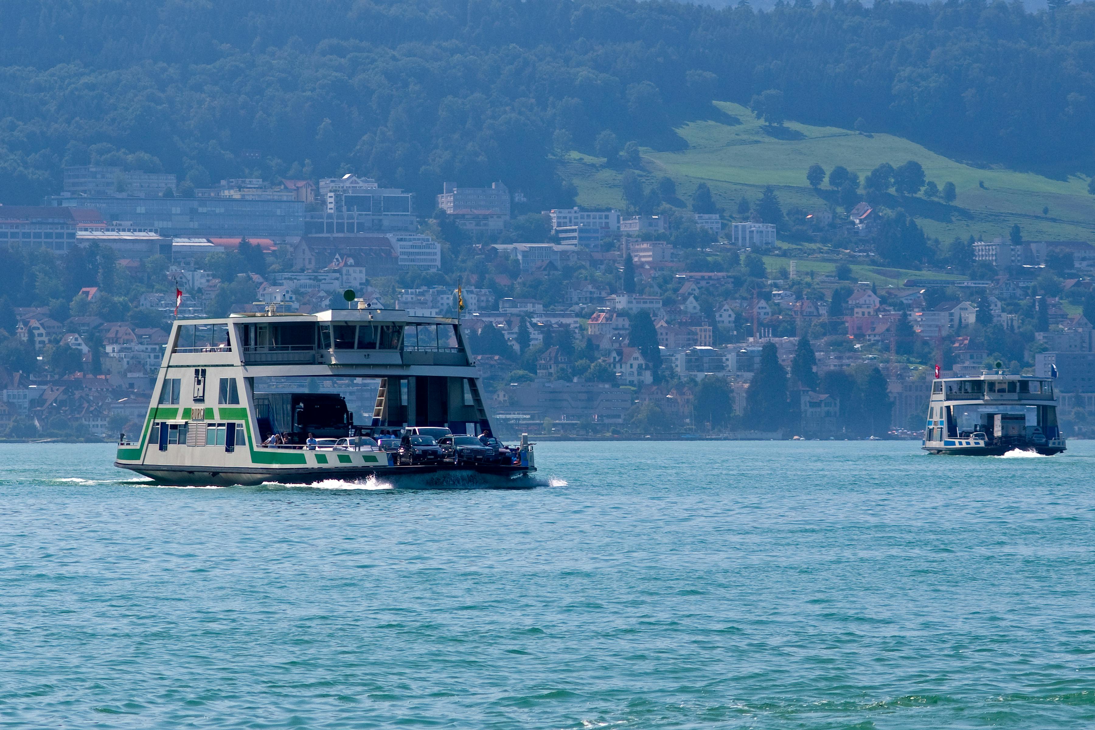 Trajekt s vozidly projíždí jezerem s kopcovitým městem a hustou zelení v pozadí.