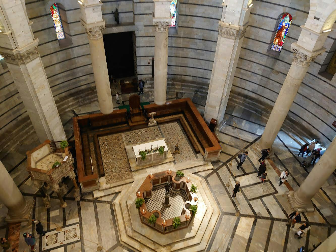 アーチ型の窓と3つの大きな扉を備えた壮大なロマネスク様式の建物。 群衆が芝生に集まり、ピサの斜塔が建物の後ろに見えます。