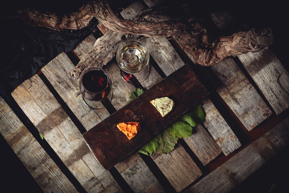 Two glasses of wine and two cheese pieces on a wooden board, set on a rustic wooden surface with a branch overhead.