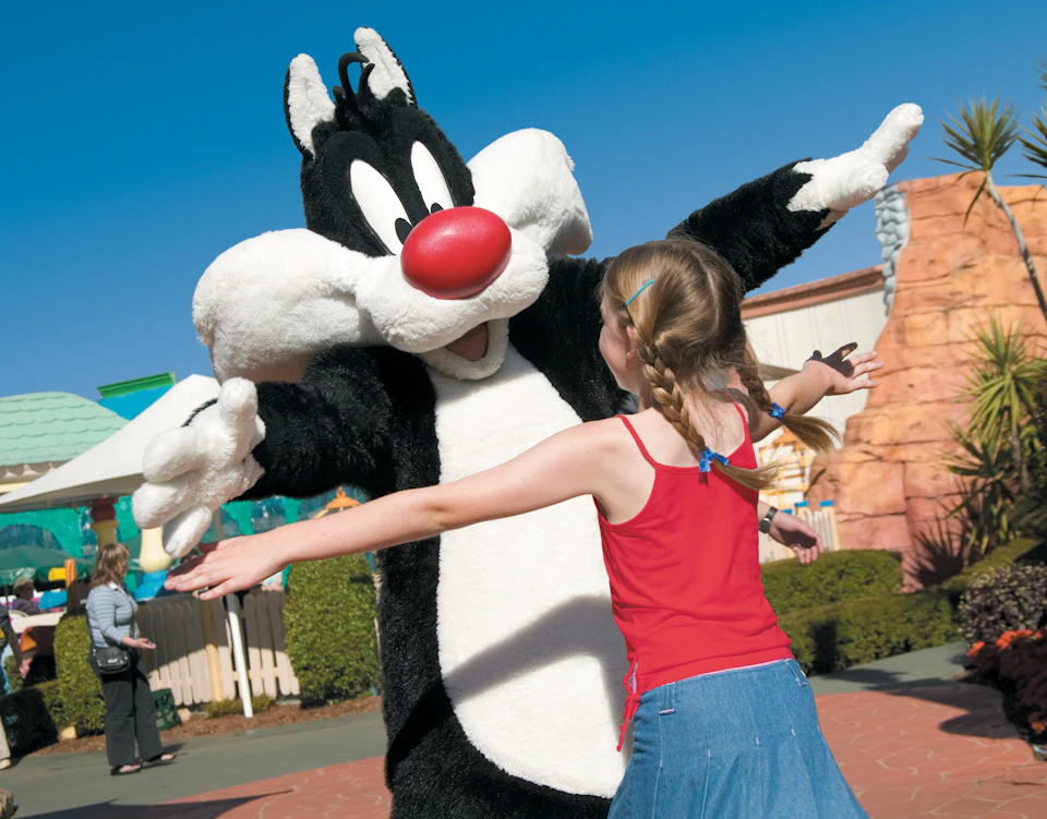 A person in a Sylvester the Cat costume extends their arms to embrace a young girl in a red tank top and blue skirt.