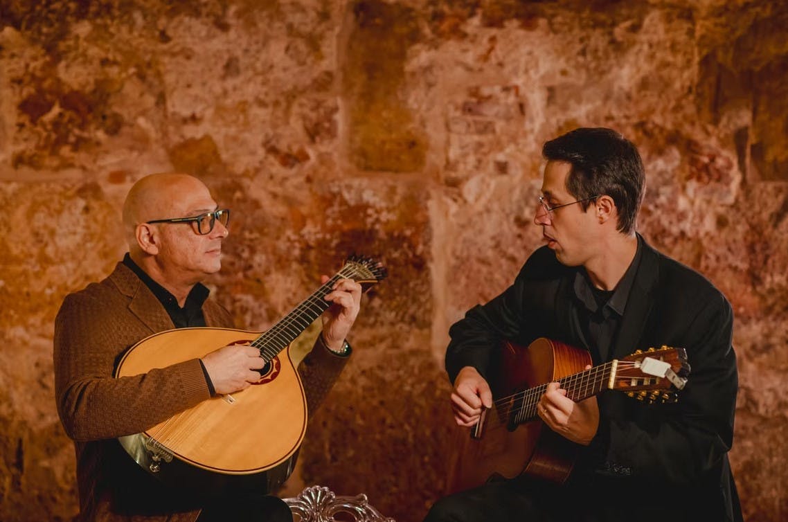 Two men seated, playing stringed instruments against a textured, rustic wall background, both wearing glasses and formal attire.