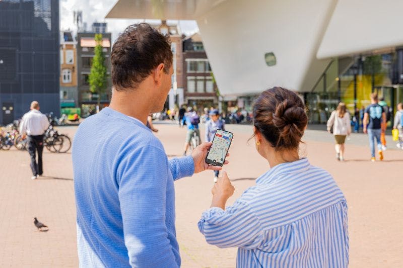 Two people outdoors looking at a phone screen displaying a map. Bicycles and buildings are in the background.