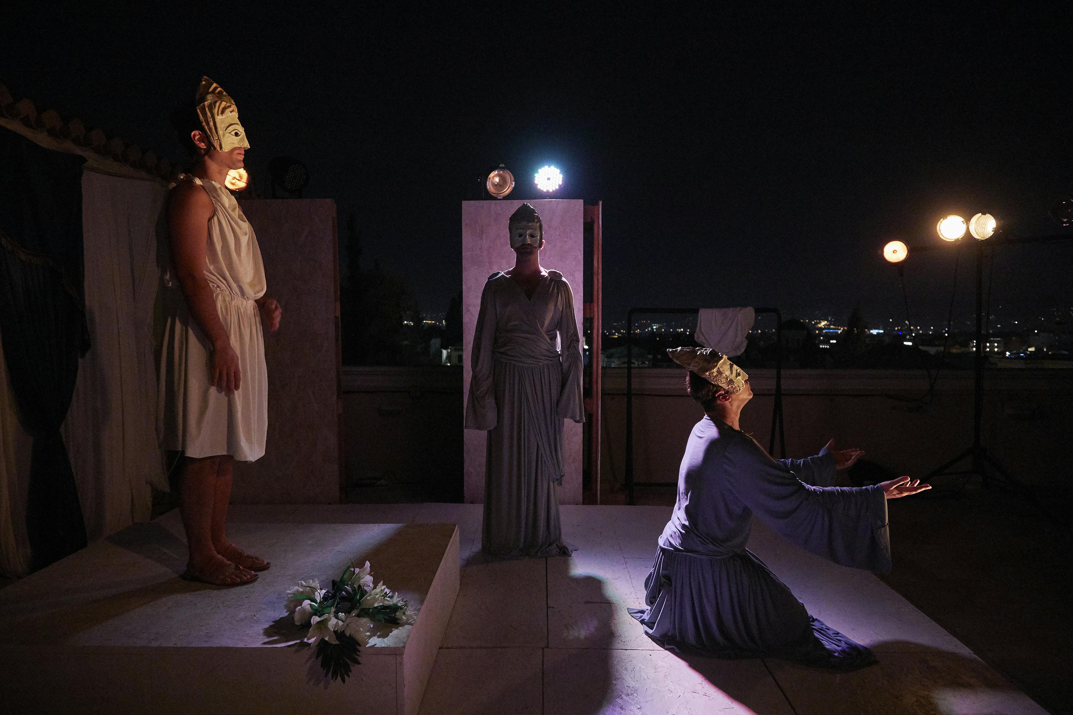 三个戴着面具的演员晚上在舞台上表演一个戏剧性的场景；一个跪着，两个站着。 道具包括鲜花和洗衣架。