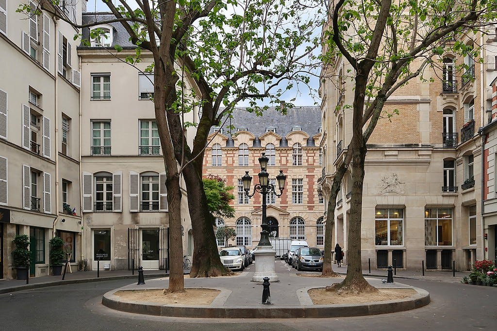 Place urbaine bordée d'arbres avec un lampadaire central, entourée de voitures garées et de bâtiments classiques à plusieurs étages dans une lumière douce.