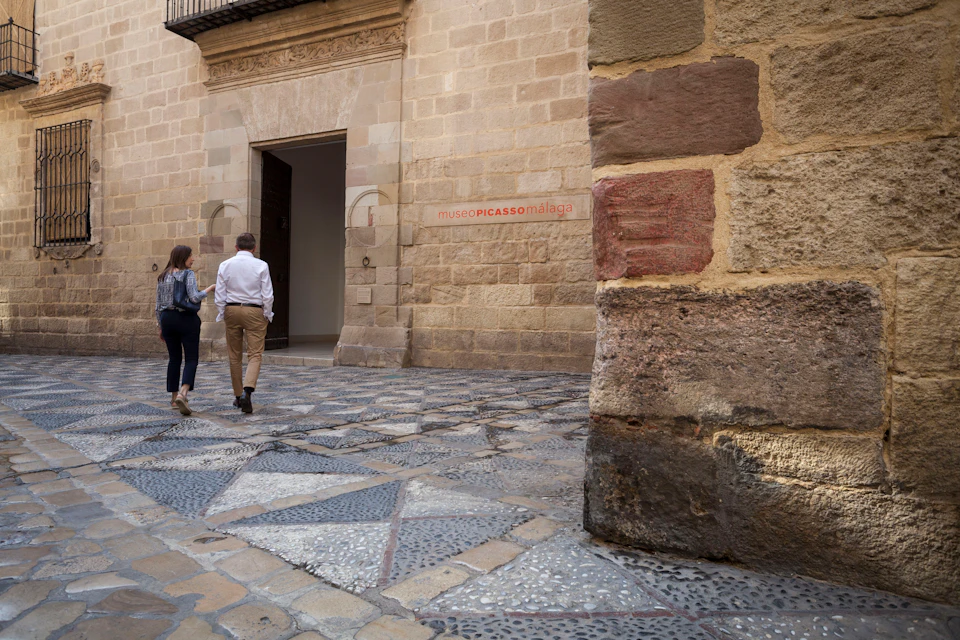 Two people walk towards the entrance of a building with a sign that reads "Museo Picasso Málaga." A stone wall is on the right.