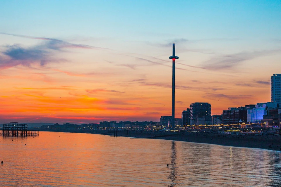 A coastal cityscape at sunset, featuring a tall observation tower and reflecting lights on the calm water.