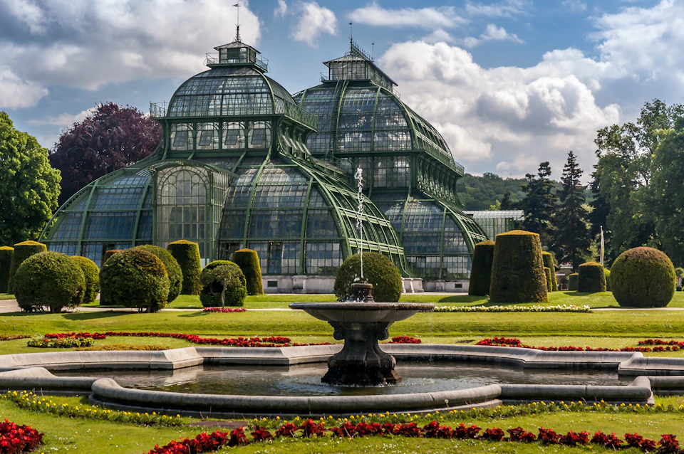 A historic greenhouse with ornate glass and ironwork, manicured lawns, topiary trees, a central fountain, and a cloudy sky backdrop.