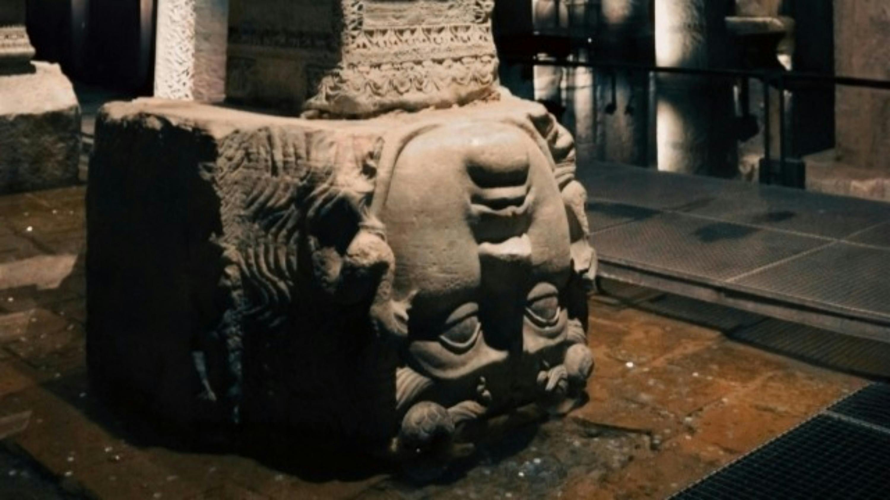 Stone column base with an upside-down carved Medusa head, illuminated, in a dimly lit historical or cultural setting.