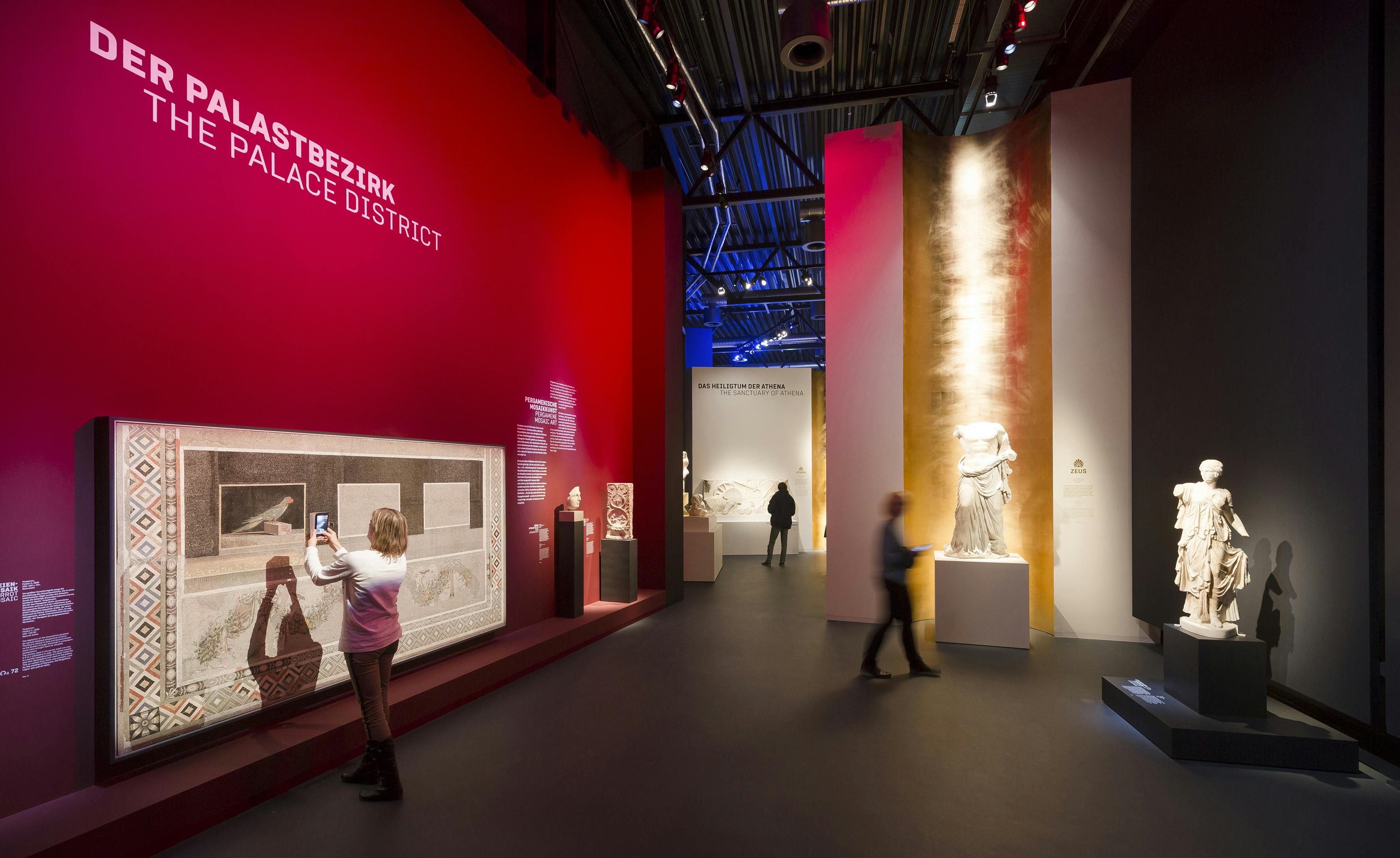 Lidé prozkoumávají uměleckou galerii s živými červenými, růžovými a zlatými stěnami a vystavují různé historické sochy a artefakty.