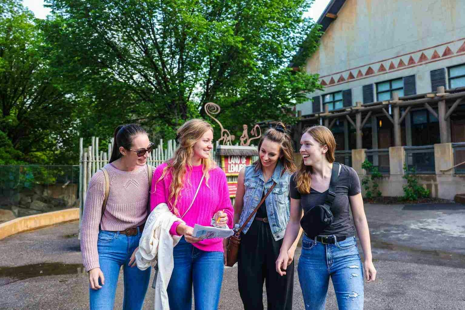 Tři ženy kráčející a usmívající se před budovou se stromem a nápisem v pozadí. Jeden drží mapu.