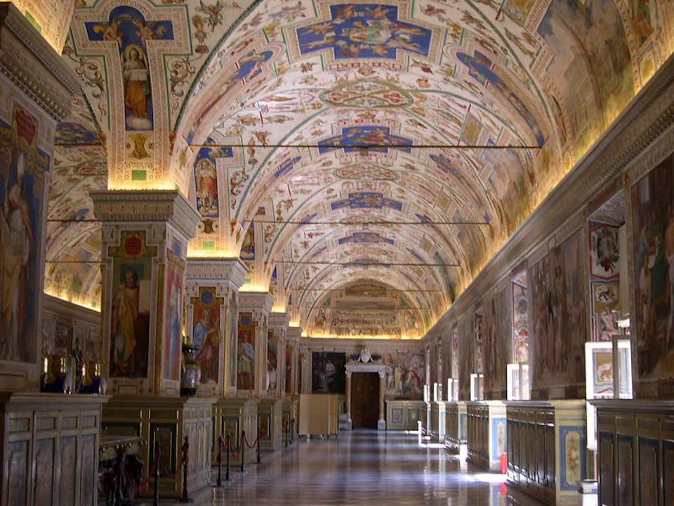 Ornate hallway with arched, intricately painted ceiling, decorated columns, wall frescoes, and a checkered floor. No people present.