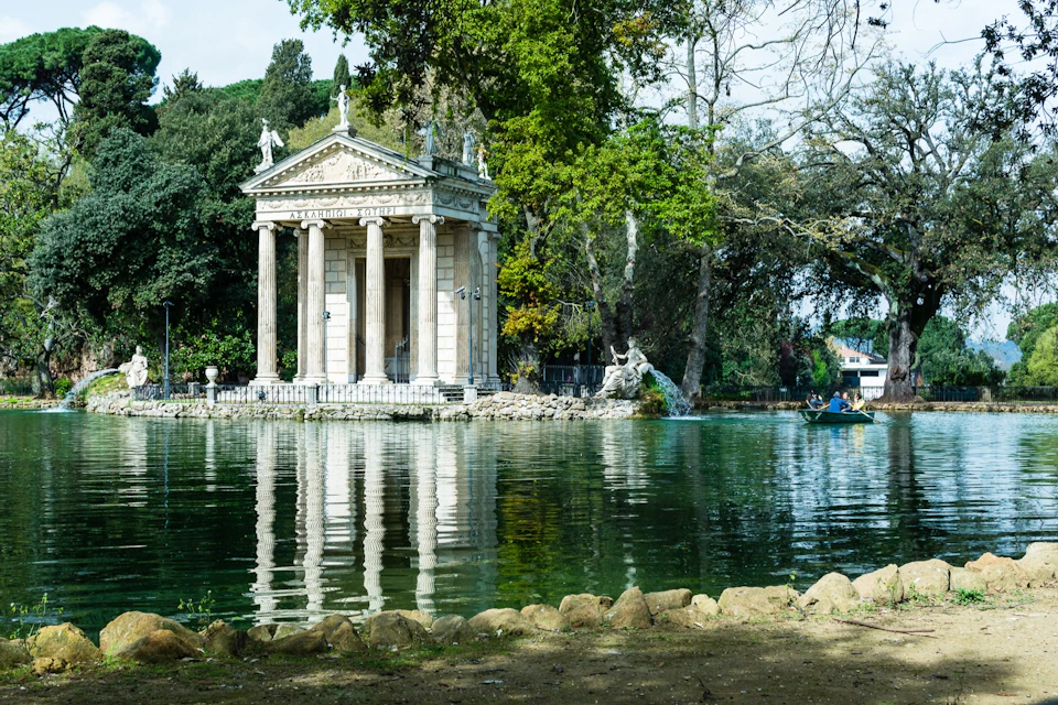 A neoclassical temple by a pond surrounded by trees, with a small boat visible on the water.