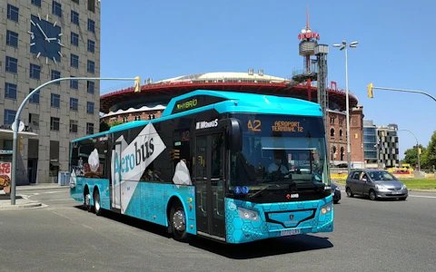 Un autobús híbrido azul rotulado "Aerobús" se dirige a la terminal del aeropuerto en una zona urbana con edificios alrededor.
