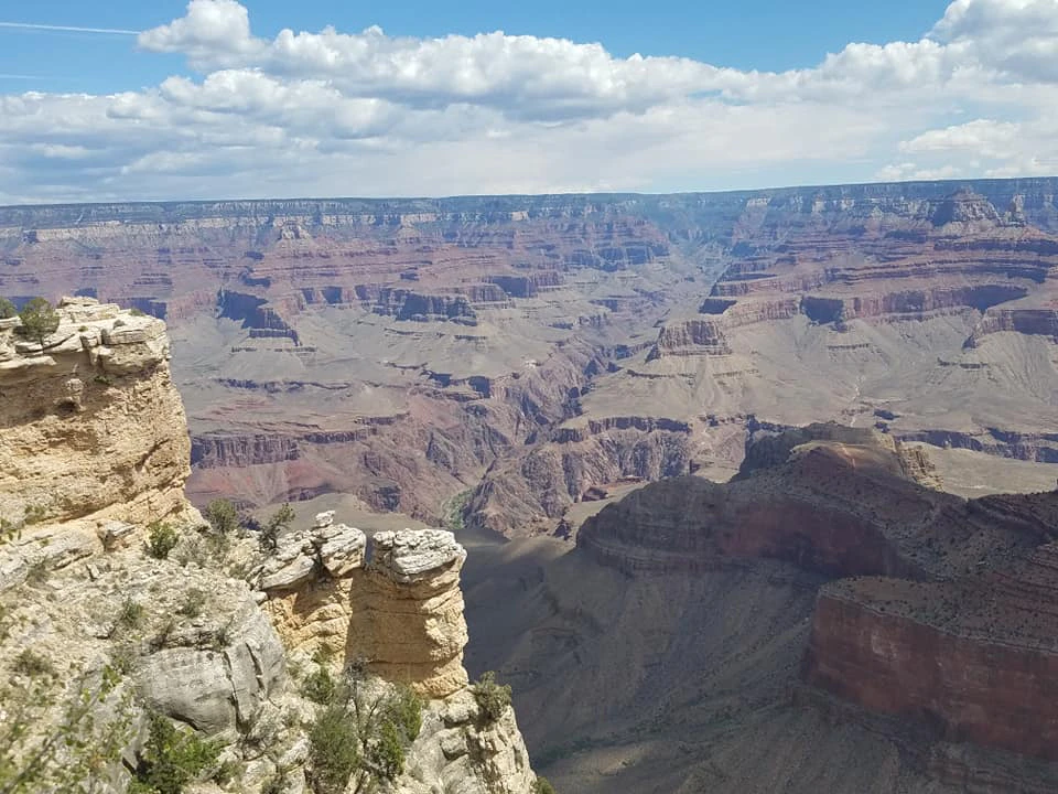 Scenic view of the Grand Canyon with layered rock formations, deep valleys, and a cloudy blue sky in the background.