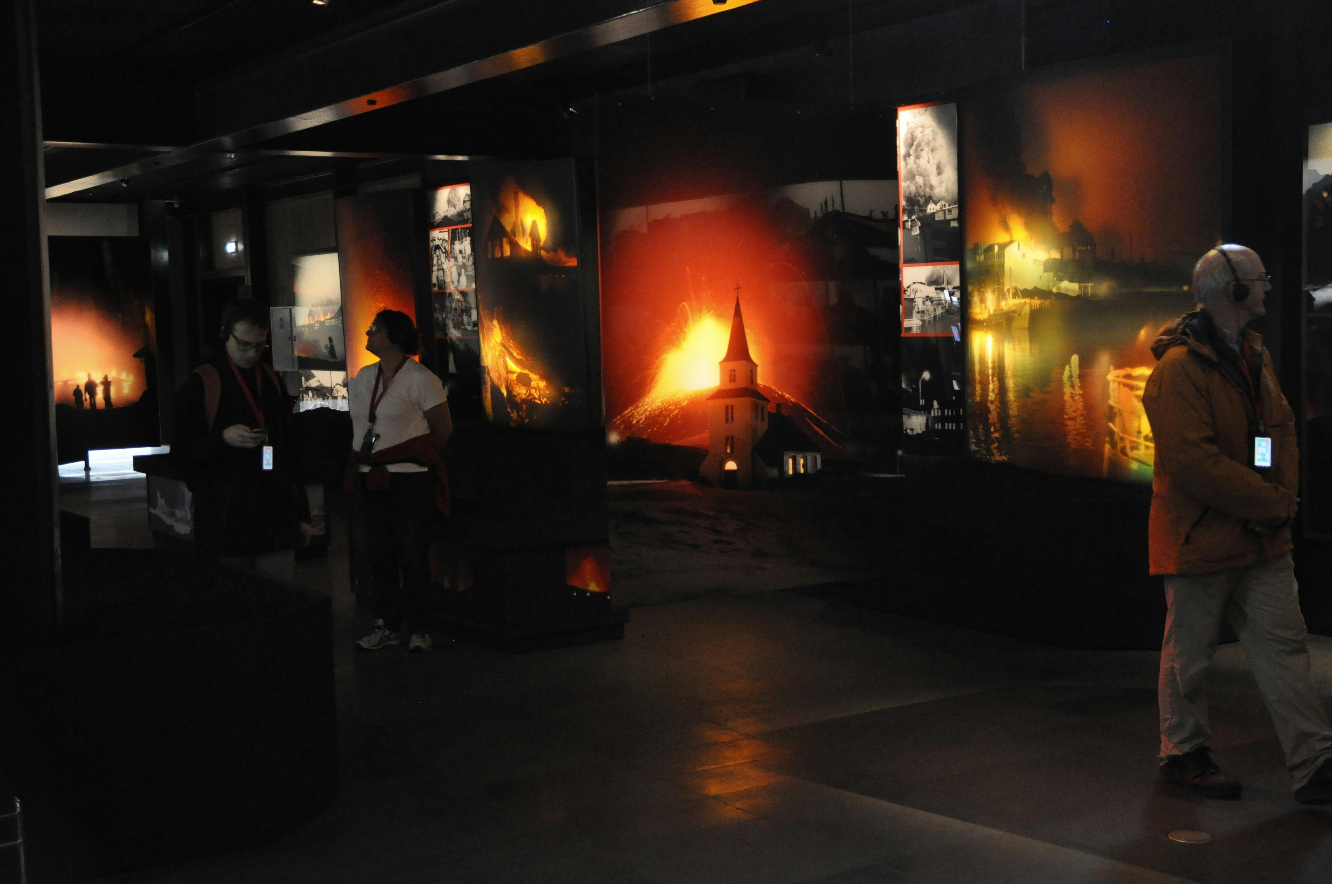 A dimly lit museum exhibit featuring images of volcanic eruptions and a church steeple. Two people observe the display.