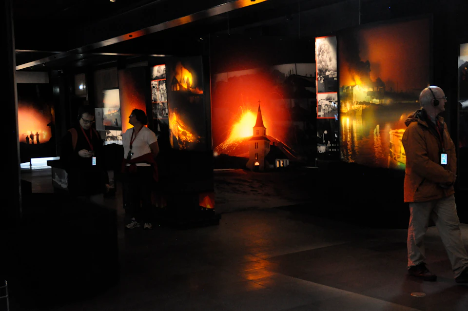 A dimly lit museum exhibit featuring images of volcanic eruptions and a church steeple. Two people observe the display.