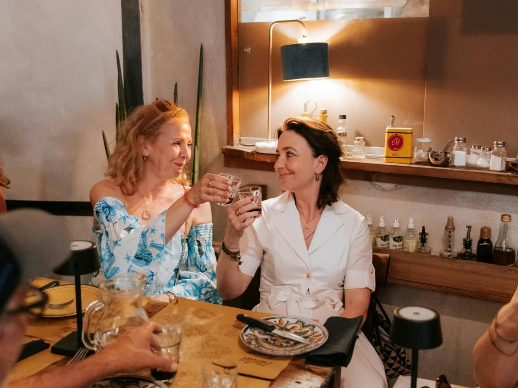 Due donne sedute a un tavolo da pranzo in casa, che brindano con i bicchieri, circondate da piatti, bicchieri e decorazioni.
