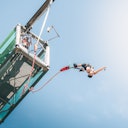 Persona che fa bungee jumping da una piattaforma con un cielo azzurro sullo sfondo. Due persone sono in piedi sulla piattaforma.