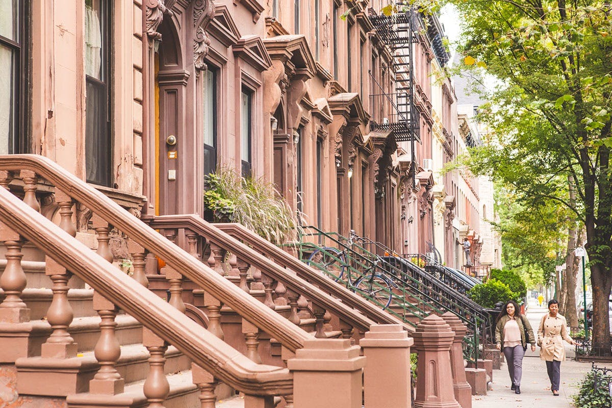 В солнечный день два человека гуляют по тротуару, выложенному коричневыми зданиями и деревьями.