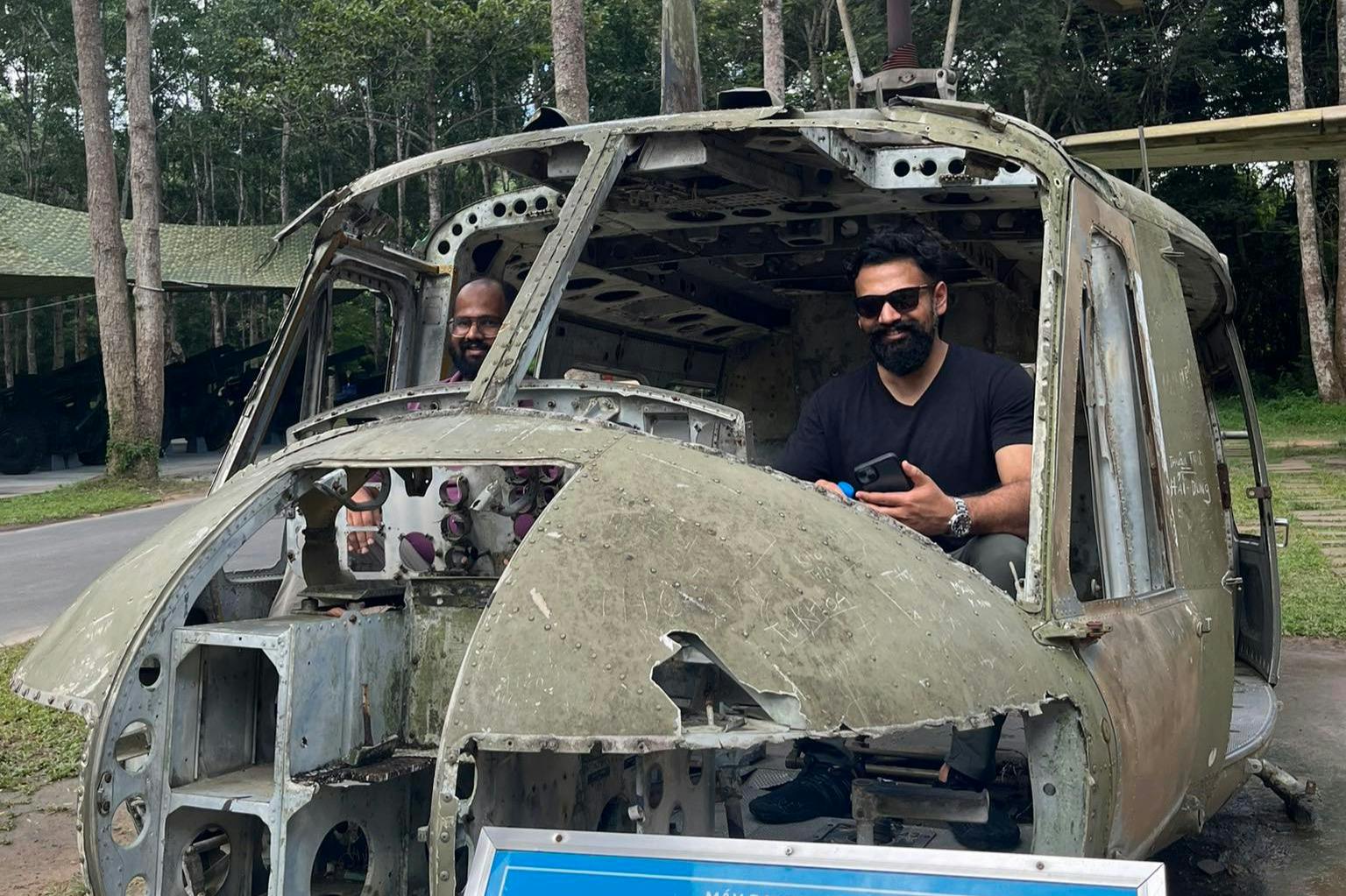 Man sitting inside a decaying, abandoned helicopter's cockpit, holding a phone, with greenery and trees in the background.