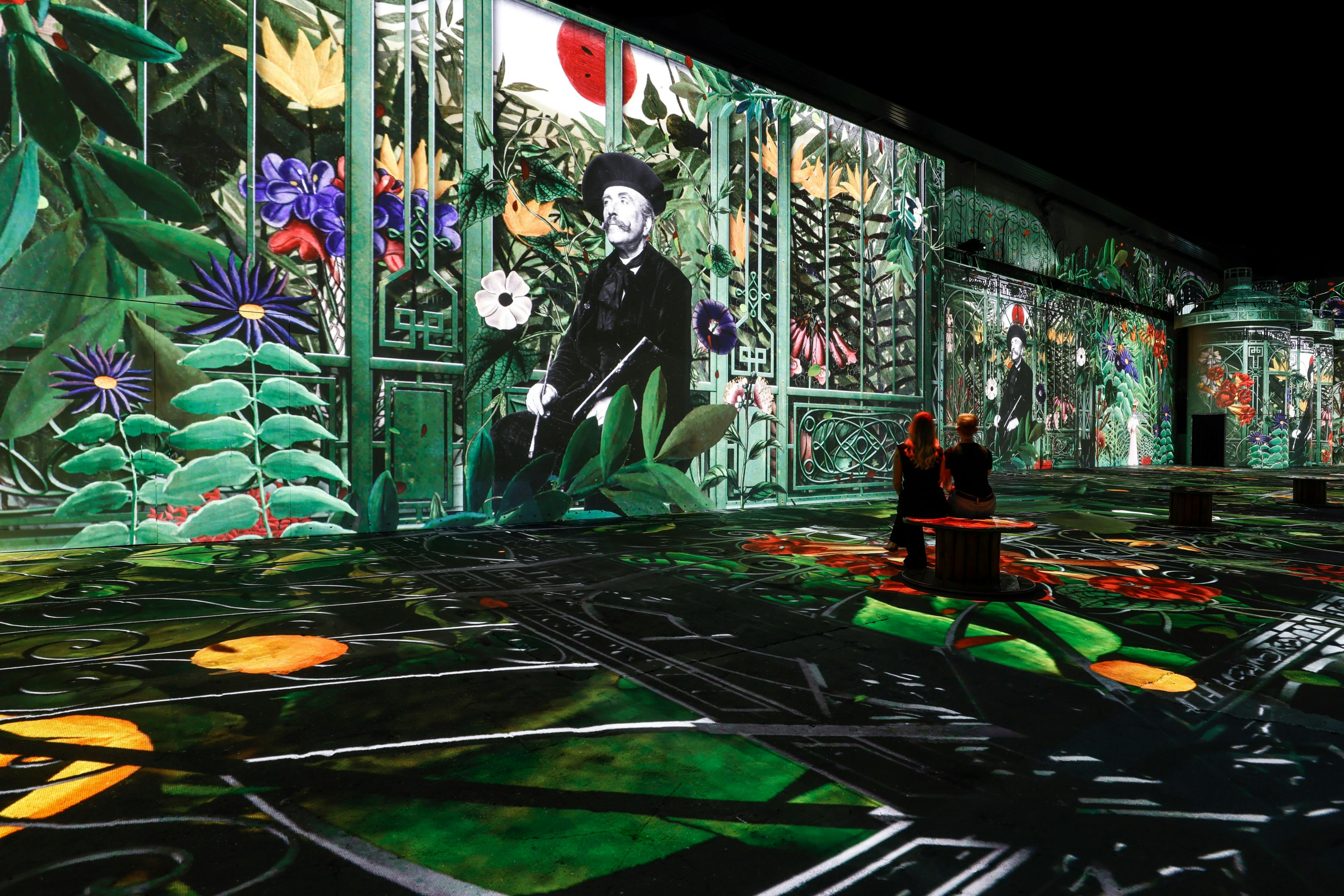 Δύο άνθρωποι παρακολουθούν μια ζωντανή ψηφιακή έκθεση τέχνης με πολύχρωμα λουλούδια και ένα ασπρόμαυρο πορτρέτο ενός άνδρα.