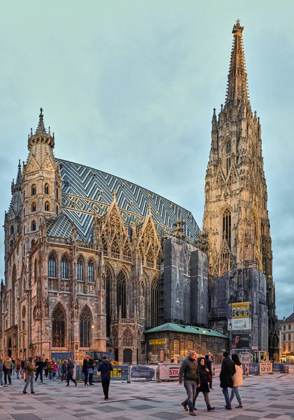 Gothic-style cathedral with a patterned roof and tall spire under a cloudy sky. People are visible near the entrance.