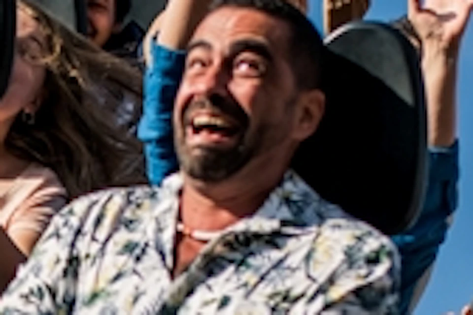 People on a rollercoaster lift their hands and scream excitedly. The ride features skulls and intricate designs. Clear blue sky above.