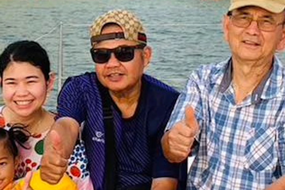 Six people, including a child, sit on a boat with a scenic view of green hills in the background, all giving thumbs up and smiling.