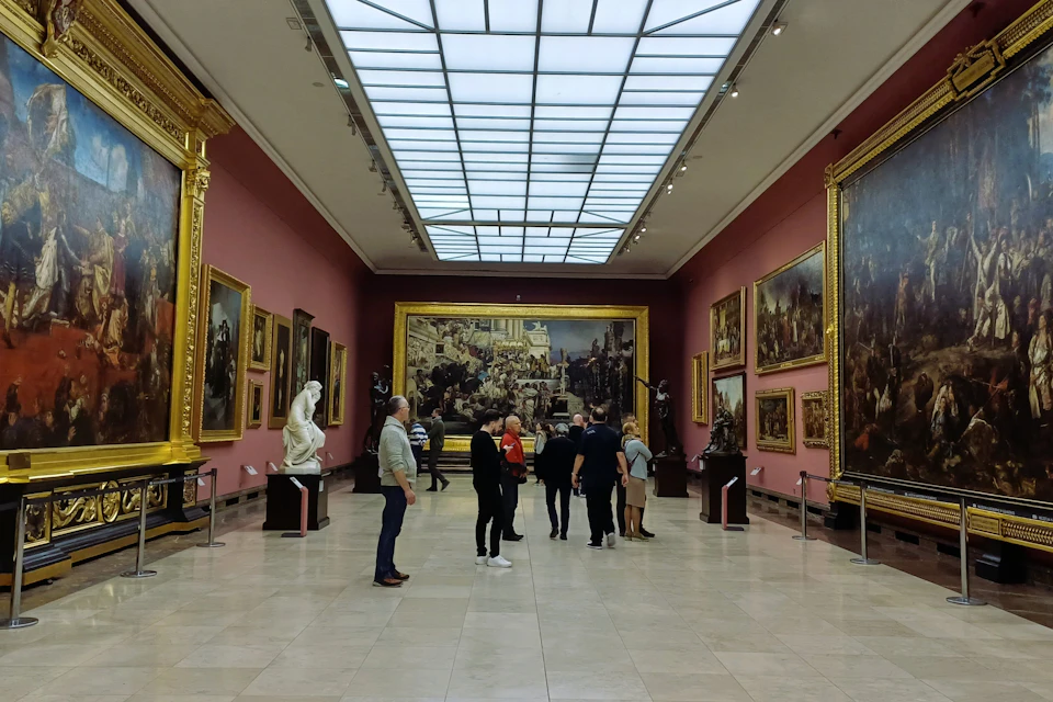Visitors observing artwork in a gallery with pink walls, large framed paintings, sculptures, and a skylight ceiling.