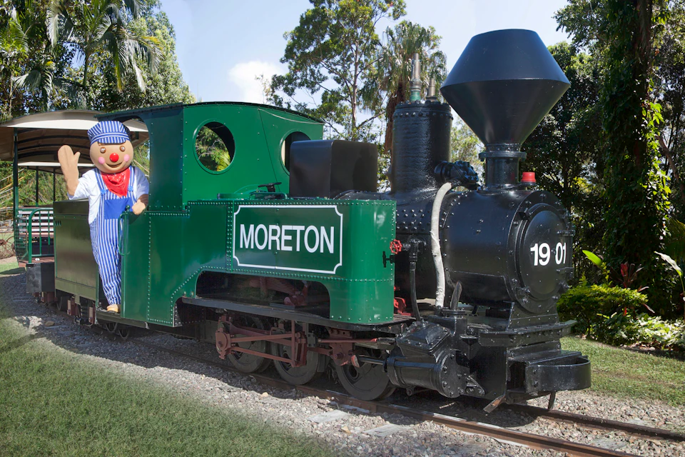 A person in a blue-and-white striped outfit and cartoonish face gestures from a green train labeled "MORETON" among trees.