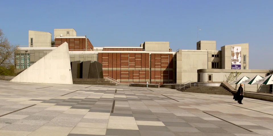 A large brick and concrete building with geometric designs, surrounded by an expansive tiled plaza under a clear blue sky.