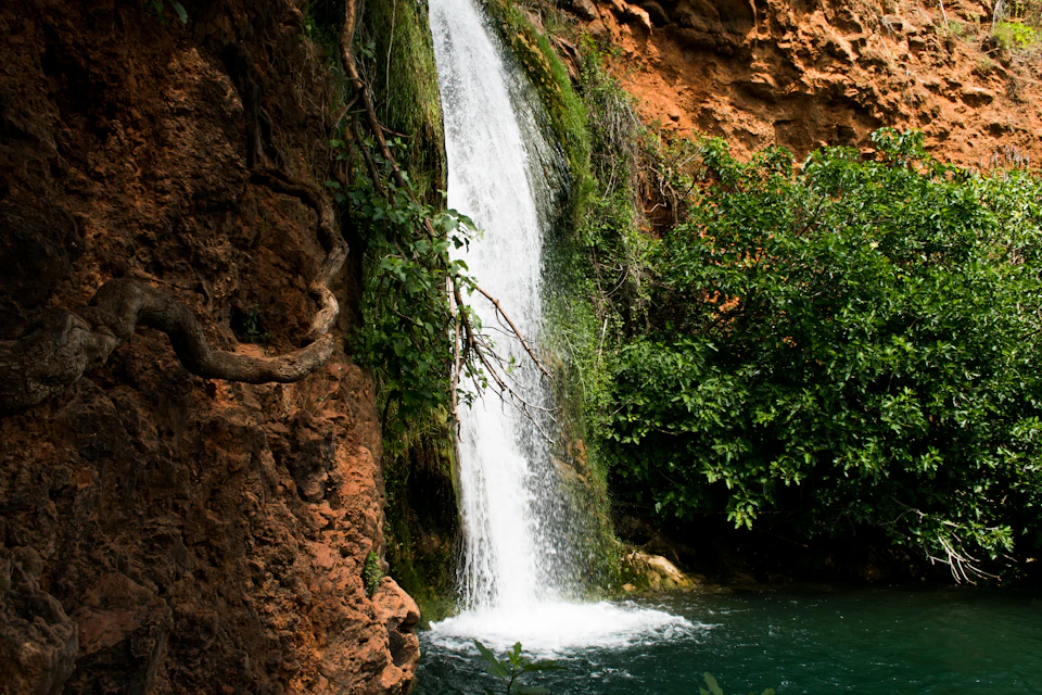 A waterfall cascades over mossy rocks into a clear, turquoise pool, surrounded by lush greenery and rugged, brown cliffs.