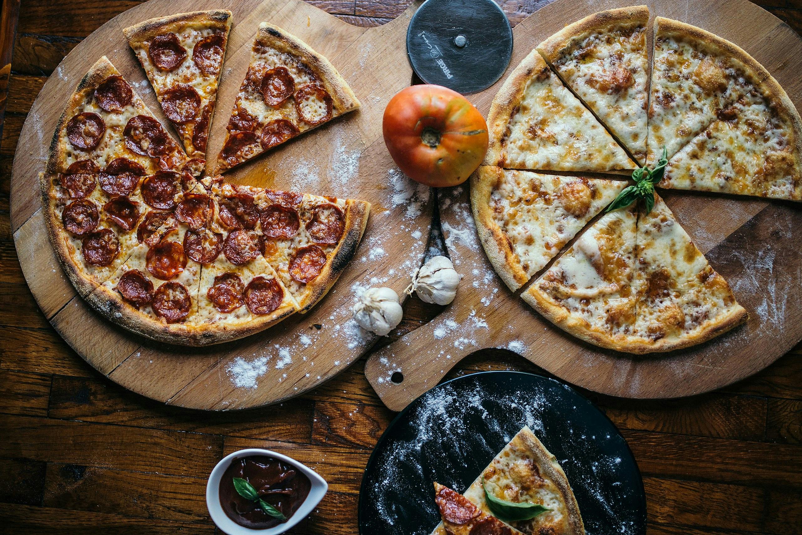 2枚のピザ、1枚のペパロニ、1枚のチーズ、トマト、ニンニク、少量のソースが入った木製のボード。