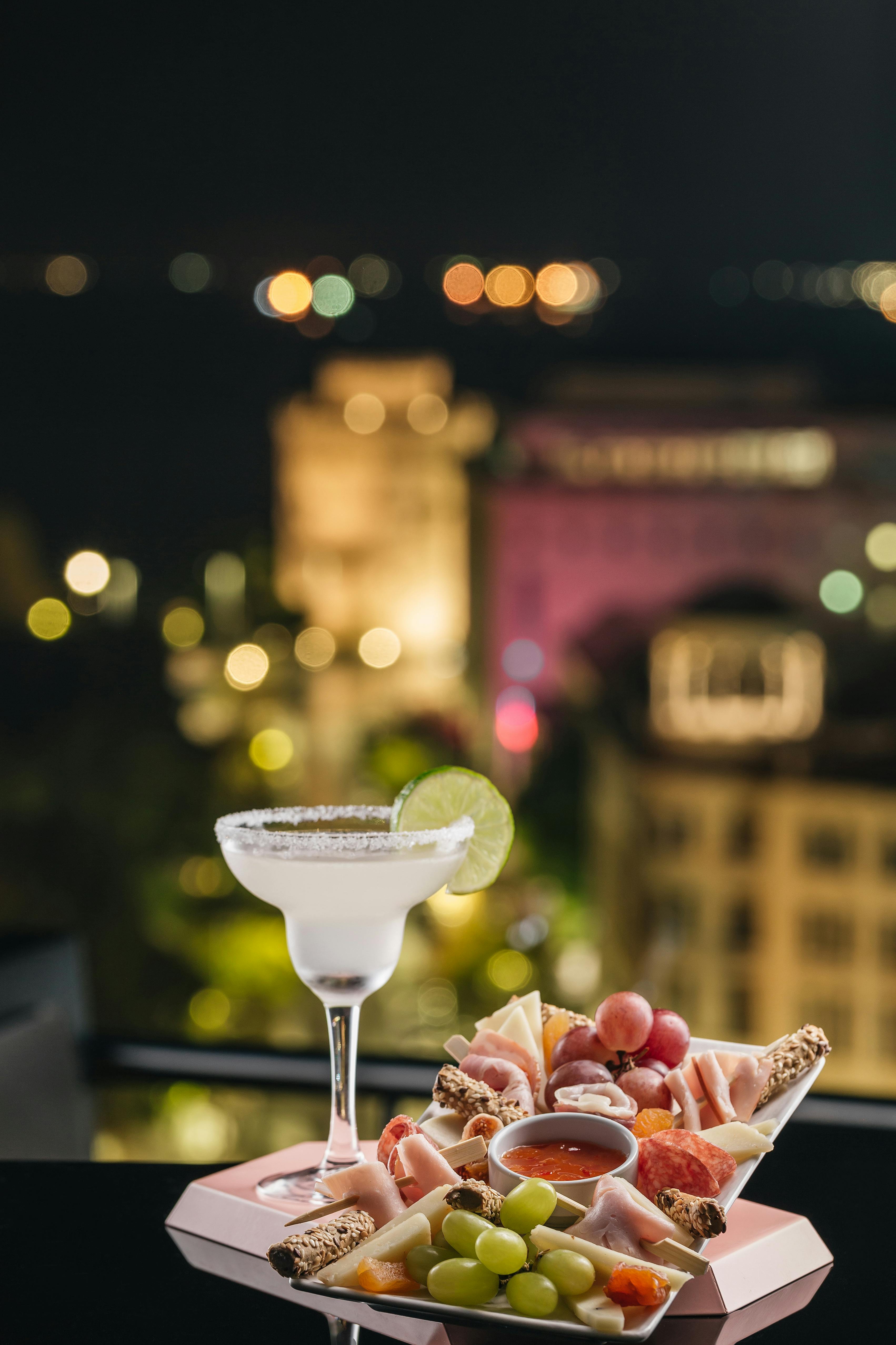 Ένα κοκτέιλ γαρνιρισμένο με μια φέτα λάιμ βρίσκεται σε ένα τραπέζι δίπλα σε ένα πιάτο με ανάμεικτα σνακ, με τα φώτα της πόλης θολά στο παρασκήνιο.