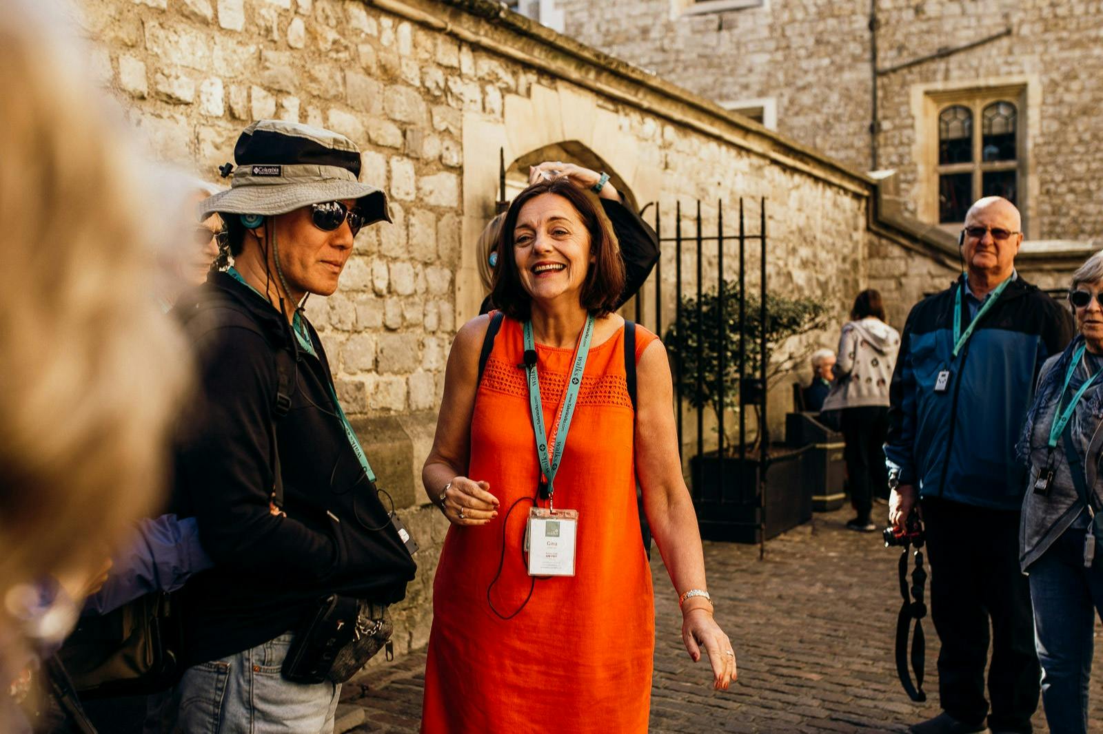 A woman in an orange dress is smiling and talking to a person wearing a hat and sunglasses on a cobblestone street by a stone wall.