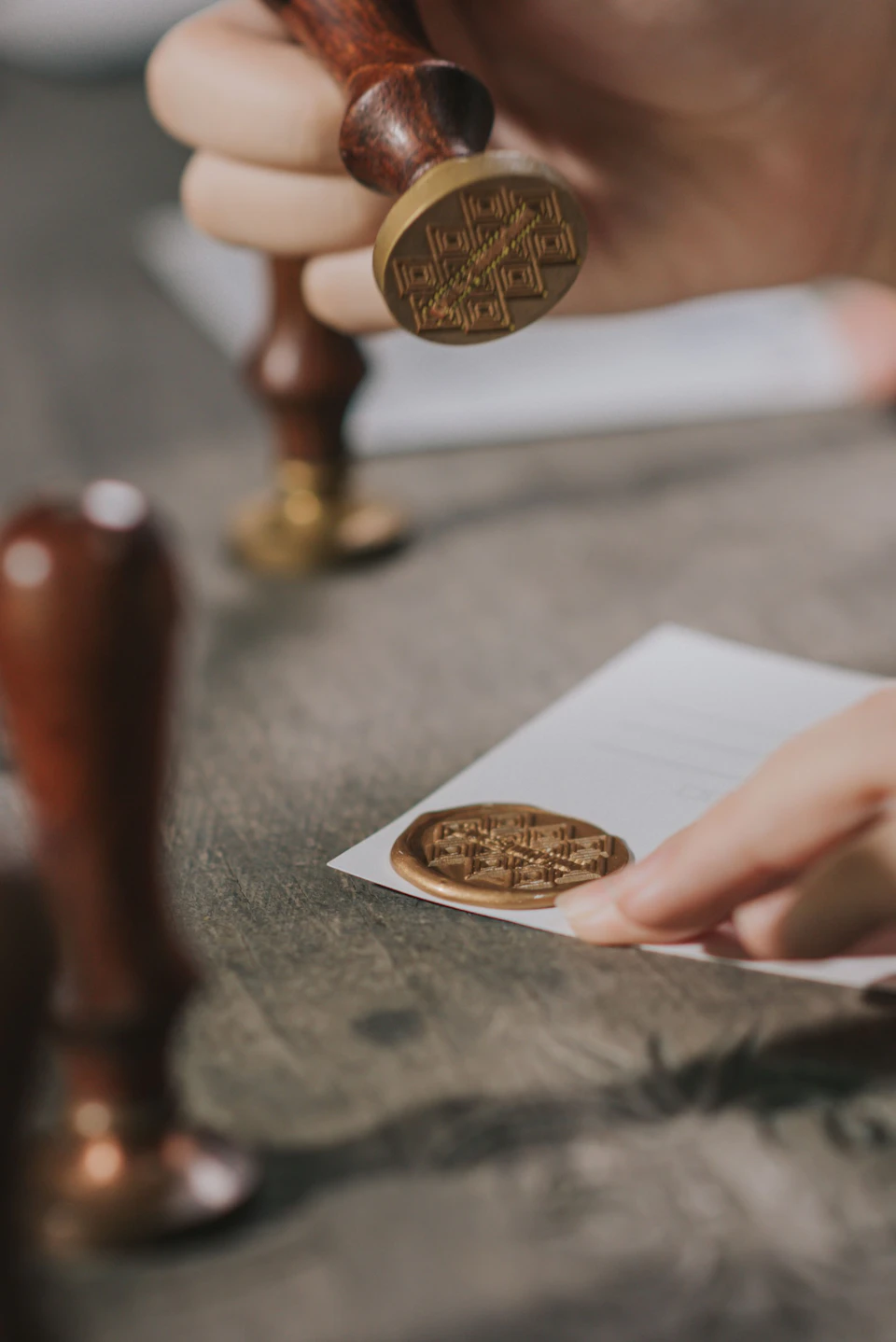 A hand pressing a wax seal onto a piece of paper on a wooden surface, with sealing tools visible nearby.