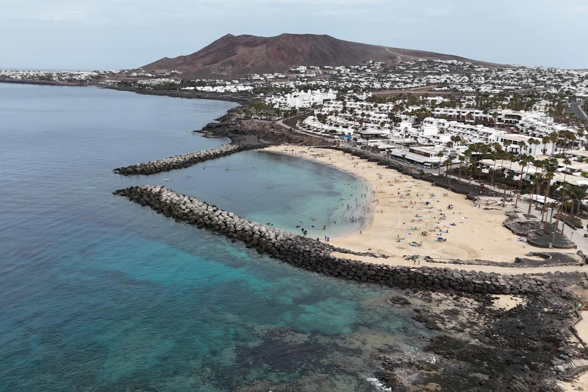 Aerial view of a beach with sand, clear water, people, and rock barriers. White buildings and a large hill are in the background.