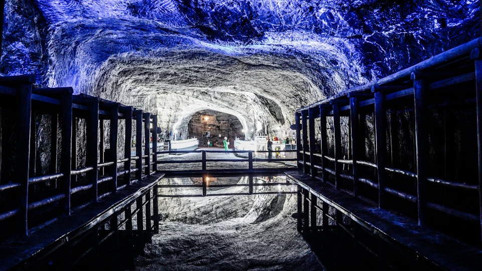 Underground cave with illuminated blue ceiling, reflective water, wooden walkway, and a person in green near a lighted area in the background.