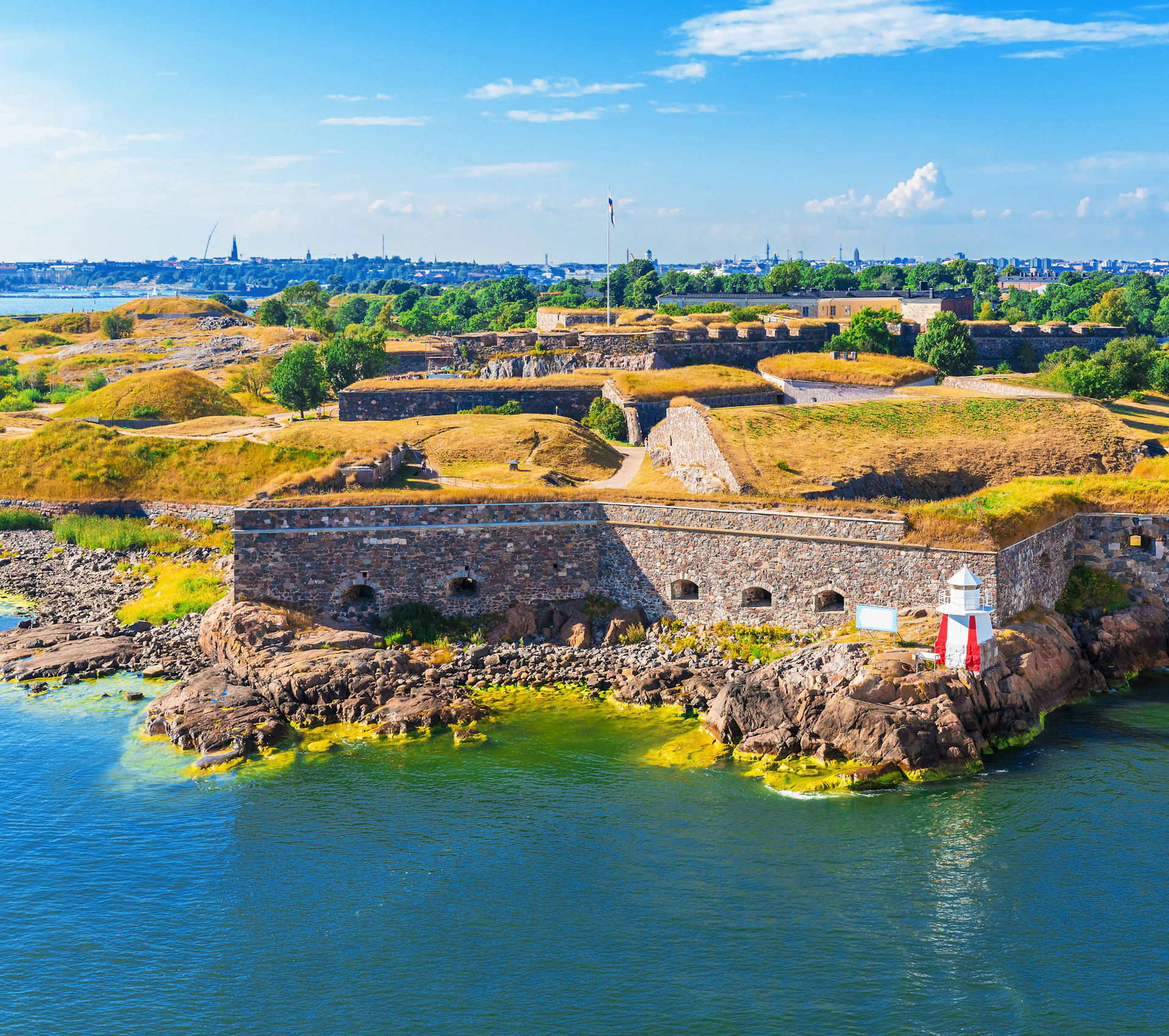 Historic fortress with stone walls surrounded by water and greenery under a blue sky with scattered clouds.