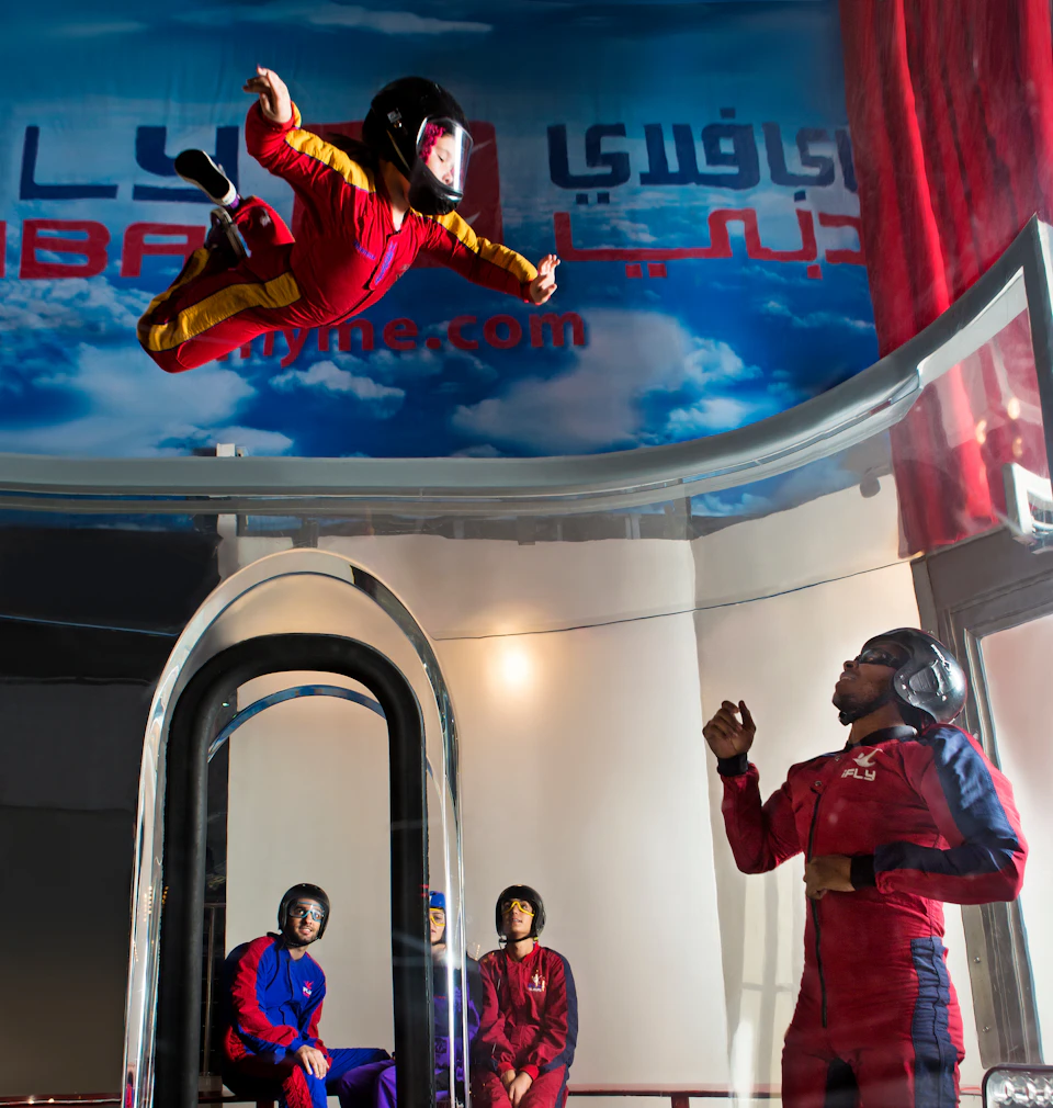 Person in red jumpsuit floating in an indoor skydiving tunnel, watched by three other people in helmets.