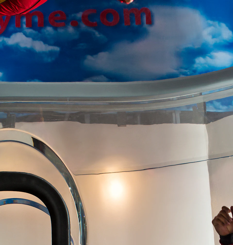 Person in red jumpsuit floating in an indoor skydiving tunnel, watched by three other people in helmets.