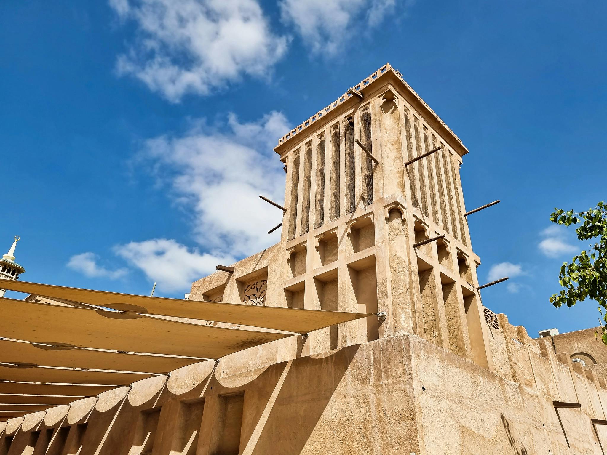 传统的米色风塔矗立在蓝天之上，云层分散，可观赏尖塔和树枝的部分景色。