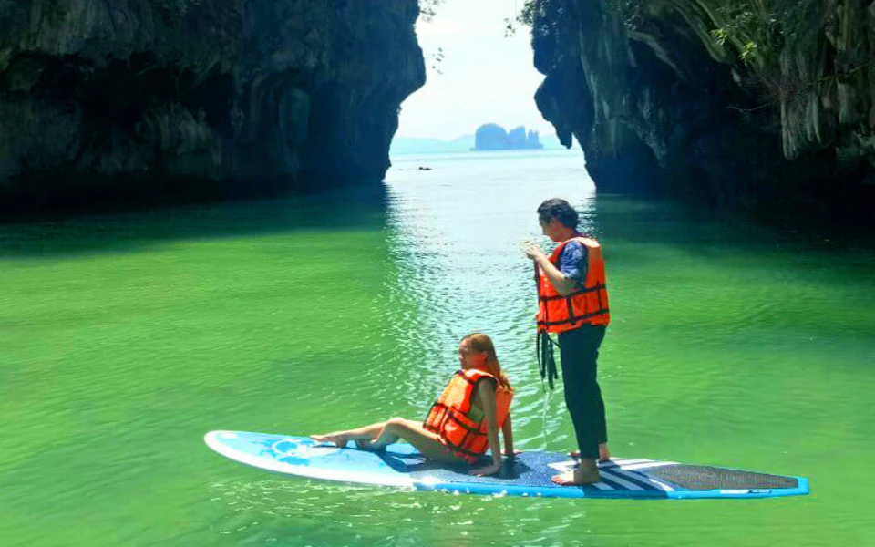 Two people in orange life vests on a paddleboard in clear green water, between rocky cliffs under daylight.