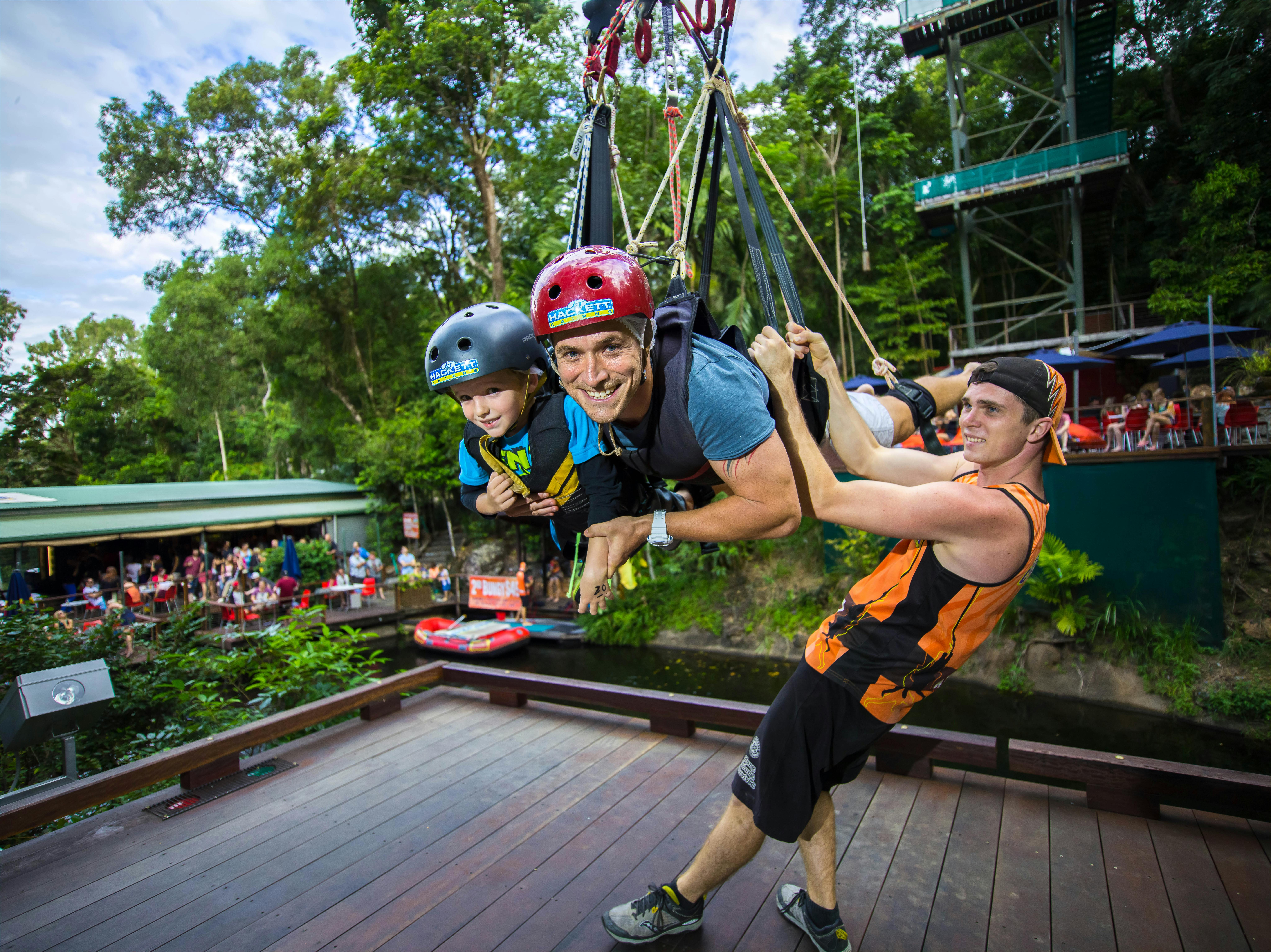 一名男子和一名儿童被绑在秋千安全带上，微笑着，而一名操作员正准备释放他们。 后面有绿色植物和人物。