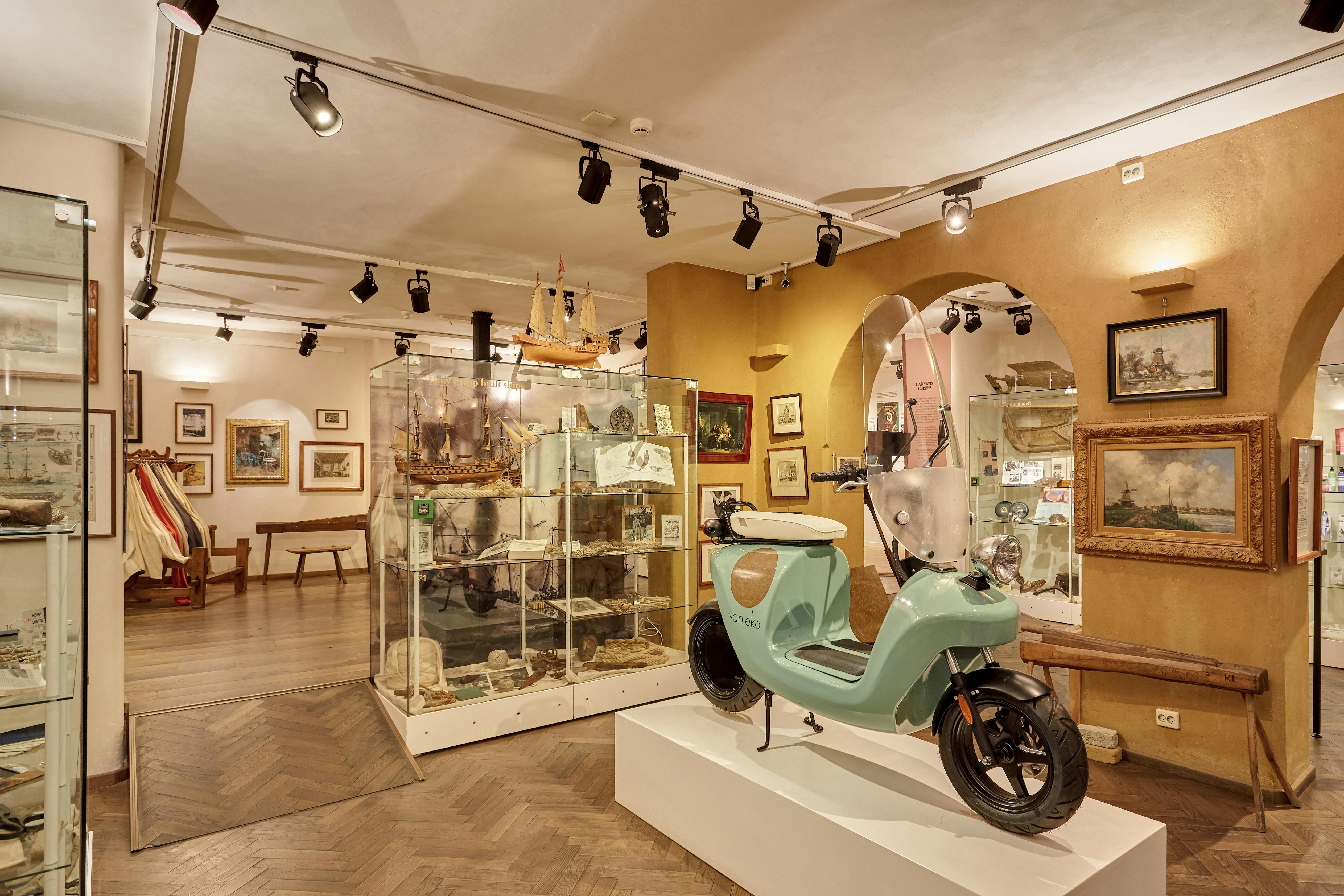 博物館の展示室では、白いプラットホームに薄緑色のスクーターが展示されており、ガラスケースには様々な工芸品や模型船が展示されています。