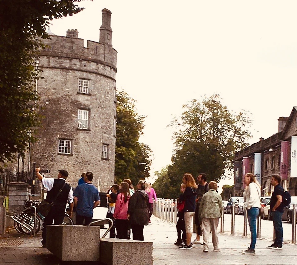 A group of people standing near a historic stone tower, with trees and a few concrete barriers in the background.
