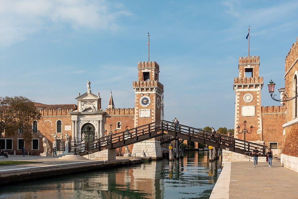 Two brick towers with clock faces, connected by a wooden bridge over a canal, with people walking nearby.
