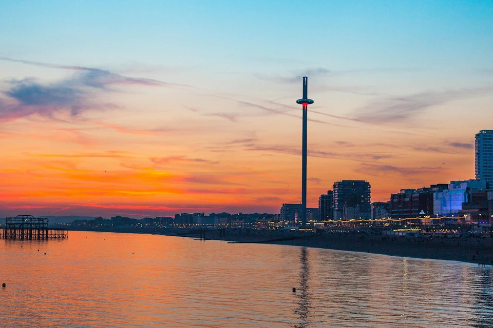A beachside cityscape at sunset, featuring a tall observation tower and illuminated buildings along the shoreline.