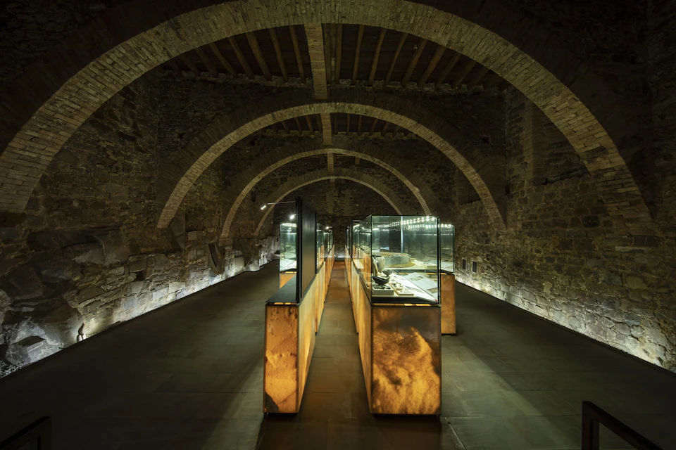 A dimly lit stone hall with arched ceilings featuring illuminated display cases with artifacts.